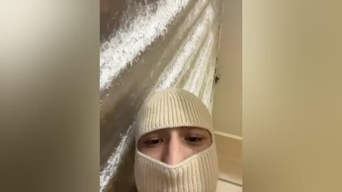 A video of a person wearing a cream-colored knit hat and matching hooded jacket, partially obscured by a silver, crinkled foil, standing in a beige hallway with a white door.