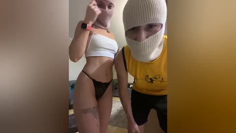 Video of two people in a dimly lit room. A woman in a white crop top and black thong stands beside a man wearing a yellow T-shirt and white mask, both with smartphones.
