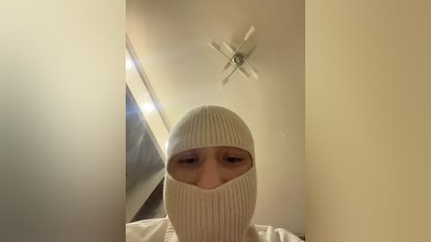 Video of a person wearing a white knit beanie and matching mask, standing in a dimly lit, beige-walled room with a ceiling fan and a rectangular window visible.