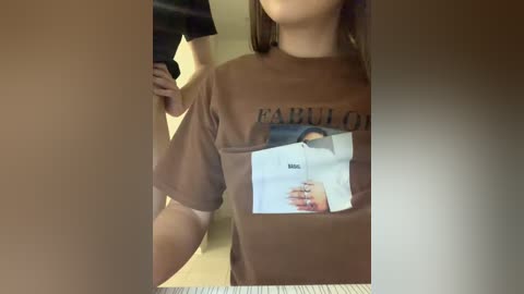 A video of a young woman with fair skin, wearing a loose brown t-shirt featuring a graphic of a man's face. Her face is partially visible. The background is blurred, with a dimly lit room.