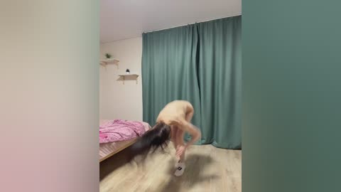 Video of a naked person with long black hair, bending over, standing on a pink bed, holding a white sock, in a teal-draped room with a wooden floor.