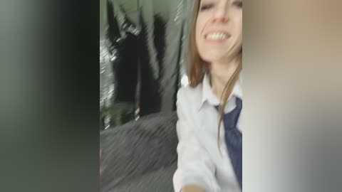 Video of a smiling young woman with light skin and long brown hair, wearing a white shirt and navy tie, standing in a blurred outdoor setting with green foliage in the background.