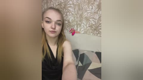 Video of a young woman with light skin and long blonde hair, wearing a black sleeveless top, sitting on a beige couch with geometric-patterned pillows. The background features a beige wall with a pattern of abstract brown and white leaves.