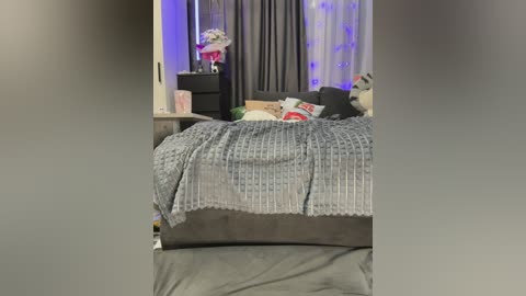 Video of a cluttered bedroom with a messy bed, gray bedding, and a gray curtain backdrop. A gray side table holds a vase with flowers.