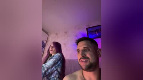 Video of a man and woman, both topless, in a dimly lit room with purple lighting. The man, with a beard, looks towards the camera, while the woman, with long purple hair, leans against a wall.