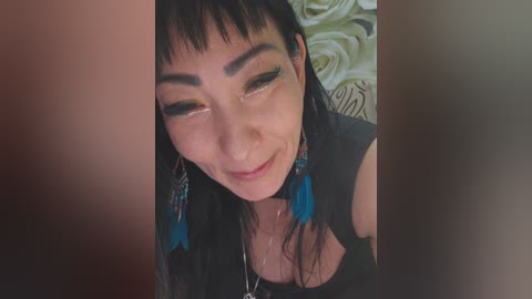 Video of a smiling, light-skinned woman with straight black hair, wearing black earrings with turquoise feathers, and a black top, against a blurred background featuring a white rose.