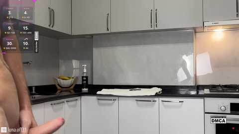 Video of a modern kitchen with white cabinets, black countertops, and a shiny stainless-steel backsplash. A person's hand is visible on the left. The right side shows a digital display with icons and \"DMCA\" text.