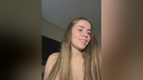 Video of a young, fair-skinned, Caucasian woman with long, straight, blonde hair, smiling, topless, indoors, with blurred background.