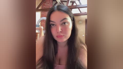 Video of a young woman with long black hair and fair skin, wearing a light pink top, indoors with a rustic wooden ceiling and brick wall background.