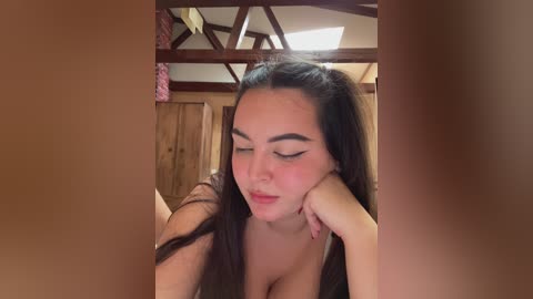 A video of a young woman with long dark hair, fair skin, and prominent eyeliner, resting her head on her hand, in a rustic wooden room with exposed beams and a ceiling light.