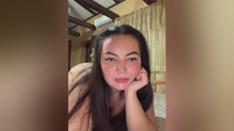 Video of a young Asian woman with long black hair, fair skin, and full lips, lying on a bed in a cozy room with wooden beams and beige curtains.