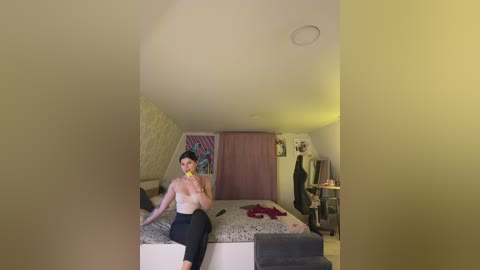 A video of a woman in a pink top and black pants, sitting on a bed with a beige blanket, in a dimly lit, cluttered bedroom with yellow walls, a desk, and a pink curtain.