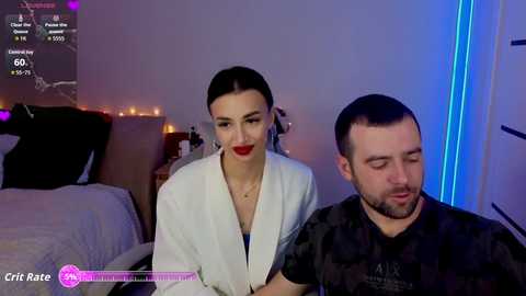 A video shows a woman with dark hair and red lipstick, wearing a white blazer, and a man with a beard, both in a dimly-lit bedroom with purple and blue lighting.
