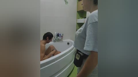Video of a small, dark-skinned man with short hair, nude, sitting in a white bathtub in a dimly lit, green-tiled bathroom, with a nurse in a white uniform standing by the door.