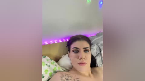 A video of a young woman with fair skin, dark hair, and striking eye makeup, lying on a bed with a green and white polka dot pillow, under a purple LED light strip.