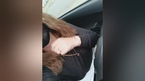 A video showing a person's shoulder and arm, wearing a black long-sleeve shirt, with a brown leather strap across the chest, seated in a car with a beige and black interior.