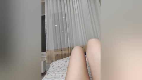 Video of a light-skinned person's legs lying on a bed, covered by sheer white curtains. Floral-patterned bedspread and a wooden headboard in the background.