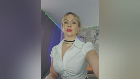 Video of a fair-skinned woman with blonde hair, wearing a white blouse and black choker, standing in a dimly lit room with blue and green LED strip lighting.