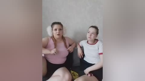 Video of two women with fair skin, wearing casual clothes, sitting side-by-side, one with braids, the other with a ponytail, in a dimly lit room.