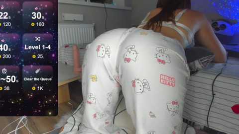 Video of a woman in white Hello Kitty pajamas, leaning over a bed in a cozy room, with a digital game interface in the background.