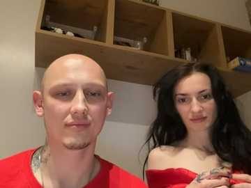 Video of a bald man with a goatee and a woman with long black hair, both wearing red shirts, standing in a room with wooden shelves and white walls.
