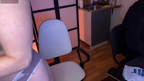 Video of a modern, minimalist room with a white chair, black desk, and wooden floor. A person in a black jacket sits at the desk, with a laptop and papers scattered around.