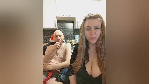 Video of a bald, shirtless man in a wheelchair eating a red lollipop, with a topless, blonde woman in a low-cut black top standing beside him in a modern kitchen.