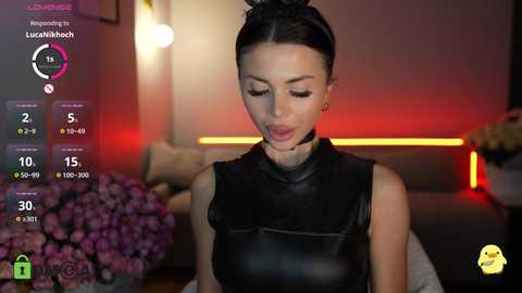 Video of a slim, dark-haired woman in a black leather sleeveless top, in a modern, dimly-lit room with a purple flower arrangement and a digital screen displaying her stats.