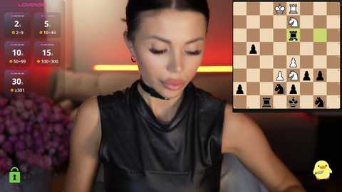 A video of a woman with dark hair, wearing a black sleeveless top, playing chess on a screen. The background is blurred, with purple flowers on the left.