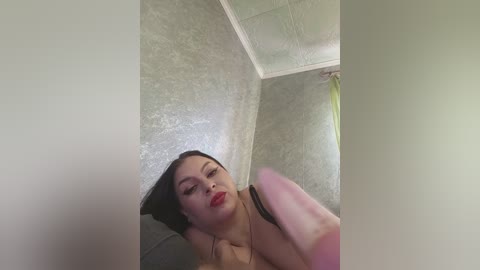 Video of a fair-skinned woman with dark hair and red lipstick, wearing a black top, leaning against a textured grey wall in a dimly lit room.