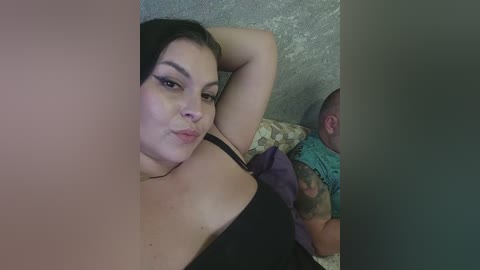 Video of a young, fair-skinned woman with dark hair, wearing a black bra, lying on a bed with a baby beside her, wearing a blue outfit.