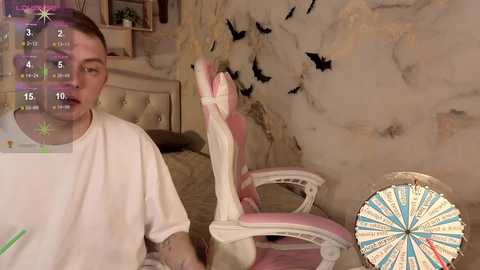 Video of a young man with short brown hair wearing a white t-shirt, sitting on a pink and white gaming chair in a cozy, dimly lit room with a beige wall and a dartboard.