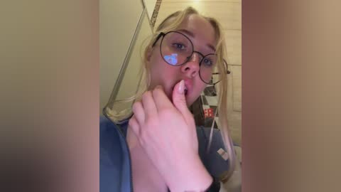 Video of a blonde woman with fair skin and large glasses, licking her fingers while wearing a blue top. She is in a bathroom with beige tiles, a mirror, and a red bottle visible in the background.
