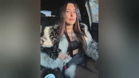 Video of a light-skinned woman with long, straight, dark hair, wearing a fur-trimmed jacket, black top, and pants, sitting in a car with a cityscape outside.