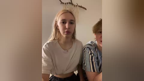 Video of a young woman with straight blonde hair, wearing a white crop top, standing indoors. A young man with short brown hair, wearing a striped shirt, is partially visible in the background.