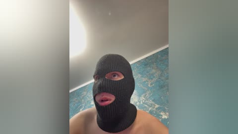 Video of a man wearing a black balaclava with eye and mouth holes, standing in a room with a blue-patterned wall and a white ceiling.