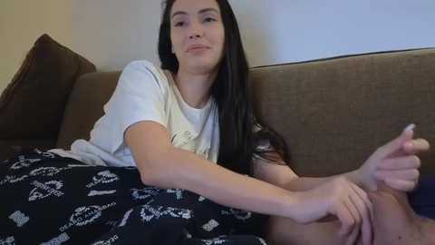 Video of a young woman with long black hair, wearing a white t-shirt and black pajamas, lying on a brown couch, holding her foot.