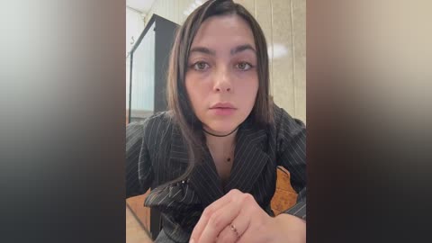 Video of a young woman with straight, dark hair, wearing a black pinstriped shirt, seated indoors with a neutral expression.