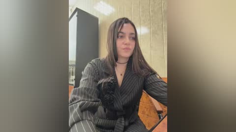 Video of a young woman with long brown hair, wearing a black pinstriped dress and black choker, seated on a wooden bench with a beige, textured wall in the background.