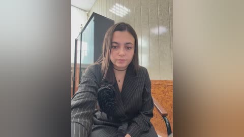 Video of a young woman with medium skin tone, straight brown hair, and a black choker, wearing a pinstripe suit, sitting on a wooden chair in a dimly lit room with wooden paneling and a TV in the background.