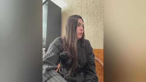 Video of a woman with long, straight brown hair, wearing a pinstriped suit, sitting in a dimly lit room with beige wallpaper and wooden paneling. She looks contemplative, leaning slightly to her right.