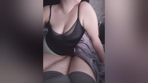 Video of a fair-skinned woman with large breasts in a black sheer bra, sitting on a bed, legs spread, wearing thigh-high gray stockings.