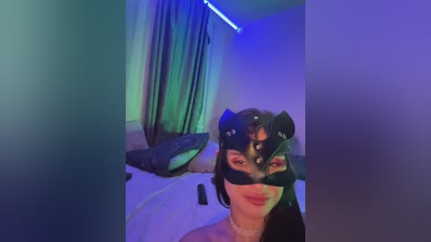 A video of a woman with light skin and dark hair wearing a black BDSM mask, standing in a dimly lit room with blue LED lighting and a bed in the background.