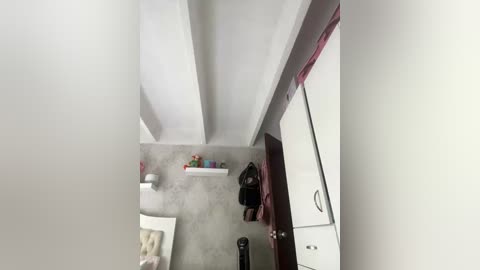 A video of a narrow, white-walled room with a gray concrete floor. A white bed with a beige blanket is on the left, a black backpack on the floor, and a red bag hangs on a white wardrobe.