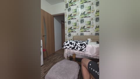 A video of a small, simple bedroom with a wooden door, a cow-print bedspread, and a striped carpet, taken from a low angle.