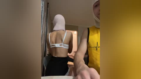 Video of a woman in a white bra and black pants, wearing a pink hijab, being restrained by a man in a yellow sleeveless shirt. The setting is a dimly lit hallway with beige walls.