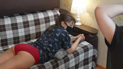 Video of a woman in glasses, wearing a dark patterned shirt and red shorts, lying face down on a plaid sofa. She's being held down by another person.