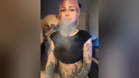 A video of a tattooed woman with pink hair, wearing a black crop top, blowing smoke into a dog's face. The background is a dimly lit room with indistinct furniture.