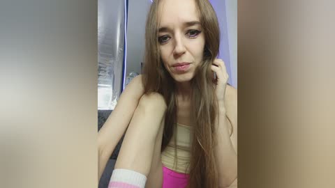 A video of a slender, pale-skinned young woman with long, straight brown hair, wearing a beige tank top and pink shorts, sitting in a modern kitchen with stainless steel appliances.