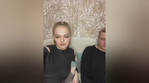 Video of two young women with pale skin, dark hair, and light makeup, sitting side by side on a couch. One wears a black top, the other a black shirt. Background features a wallpaper with gold palm trees.